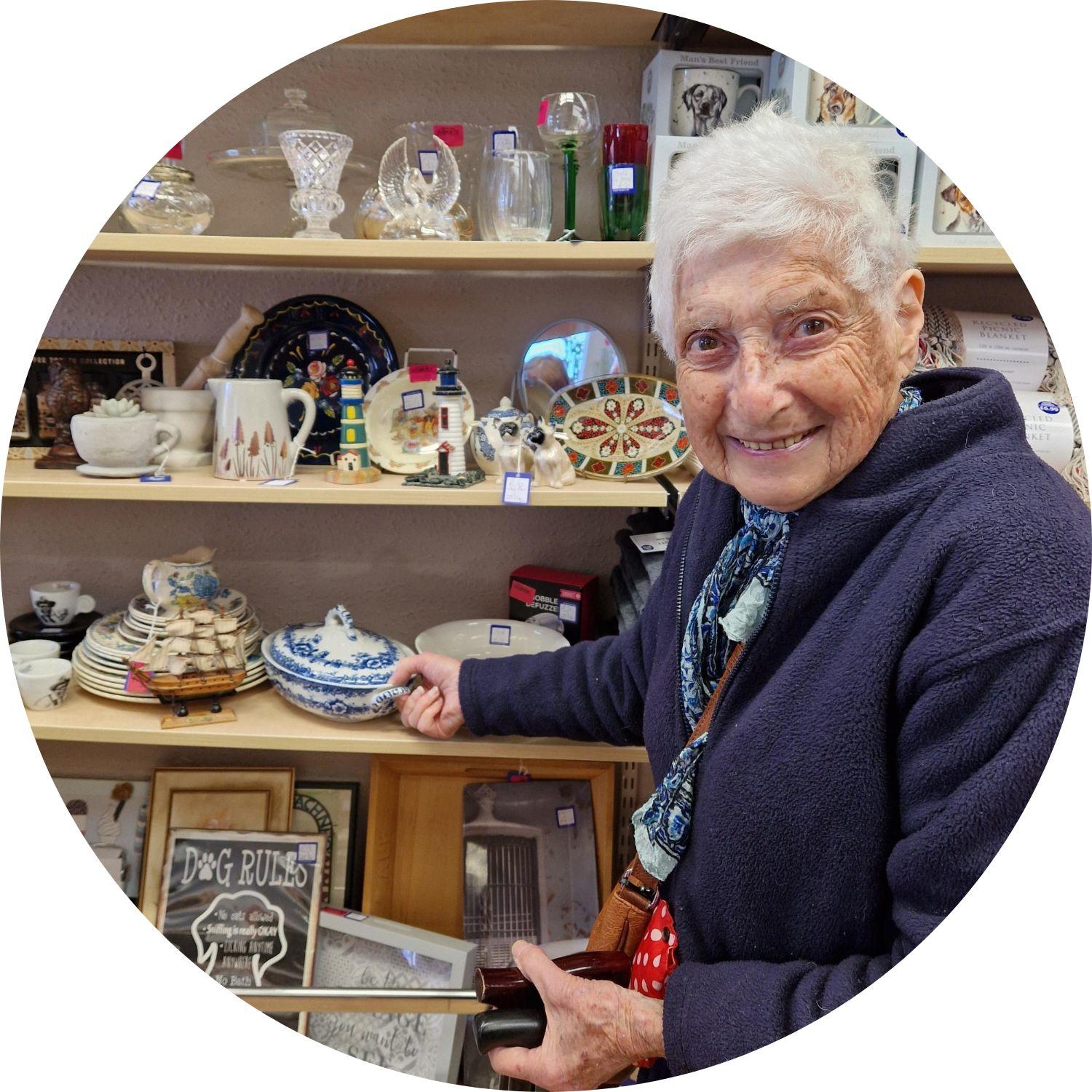 Naomi smiling and standing in front of the bric-a-brac shelf in the shop