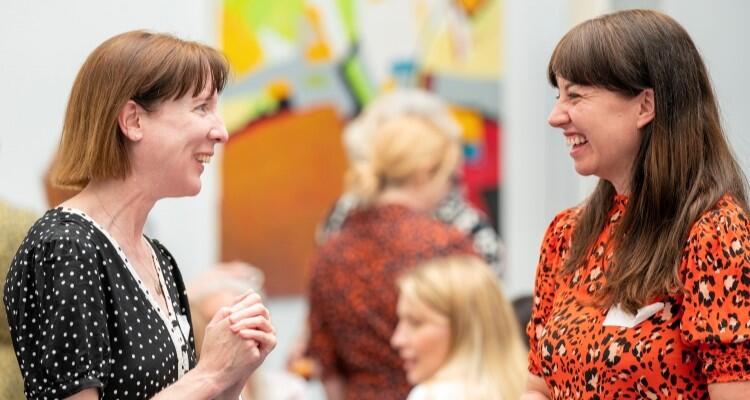 Two women standing in a room facing each other smiling.