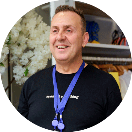 Portrait of Stojan in a charity shop looking off camera with a smile wearing two dark blue lanyards