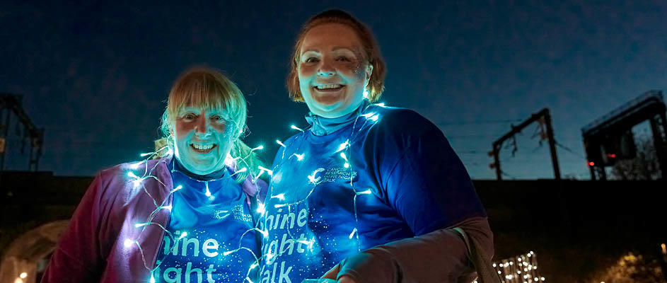 Two women smiling at the camera with fairy lights across them at Shine Night Walk