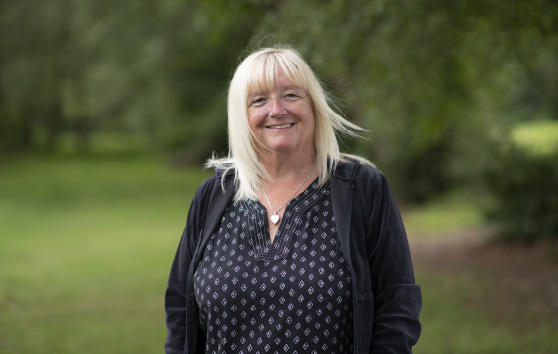 Val smiles after pledging to leave money in her Will for CRUK. Val is standing in a park a long a tree line. 