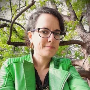 A person with dark brown hair facing the camera wearing a green jacket and glasses. They are outdoors and a leafy tree can be seen.