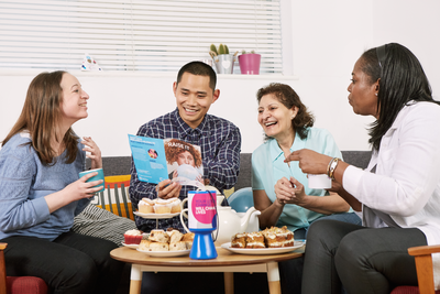 Men and women sit at a table with cakes and biscuits and are talking and laughing, they are helping at an event for Cancer Research UK and have a collection tub with them.b