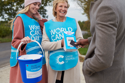 Two female volunteers with collection buckets, wearing blue World Cancer Day tabards, fundraising for World Cancer Day